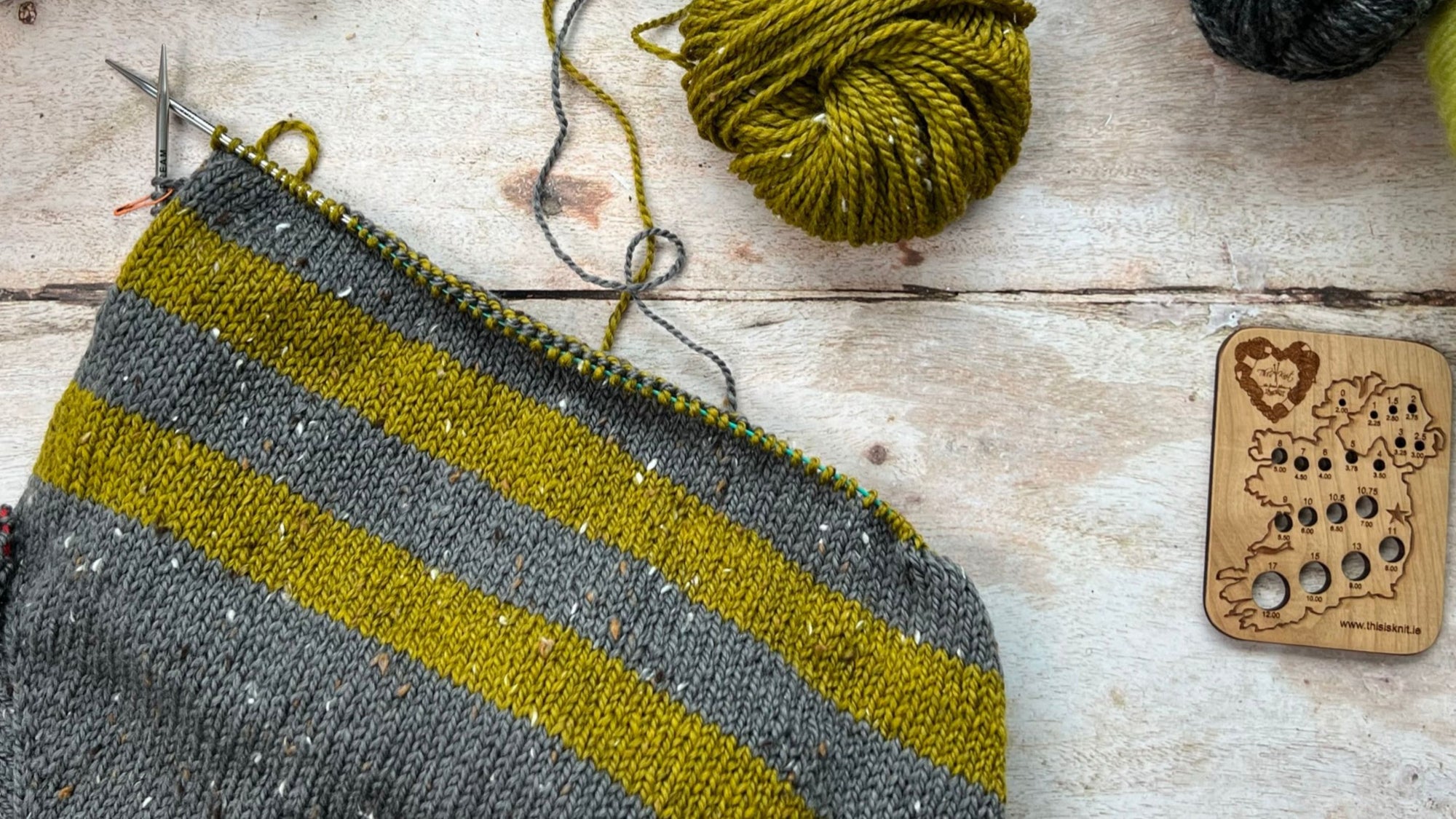 A top down view of a striped sweater in progress, knit in the round, using grey and yellow/green yarn. A needle gauge in the shape of Ireland lies to the right hand side.