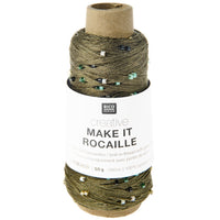 A spool of knit-in thread by Rico Design, featuring olive green yarn with small multicolored sequins, wrapped in a white paper label that reads 'creative MAKE IT ROCAILLE'.