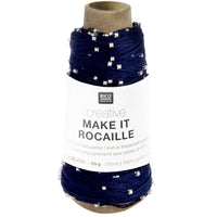 A spool of navy blue knit-in thread with small silver glass beads, wrapped in a white paper label that reads 'RICO DESIGN creative MAKE IT ROCAILLE'.